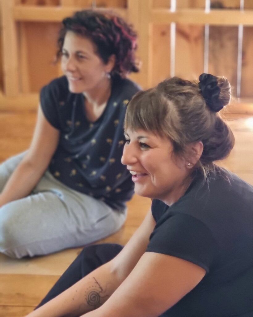Dos participantes del curso sentadas en el suelo de madera, sonriendo mientras observan un ejercicio a un lado.