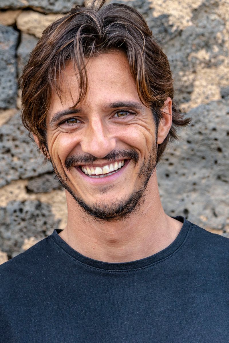 Matteo Leoni sonríe a la cámara, de medio cuerpo, con el pelo castaño ondulado y barba corta, con una camiseta oscura frente a un muro de piedra volcánica oscura.