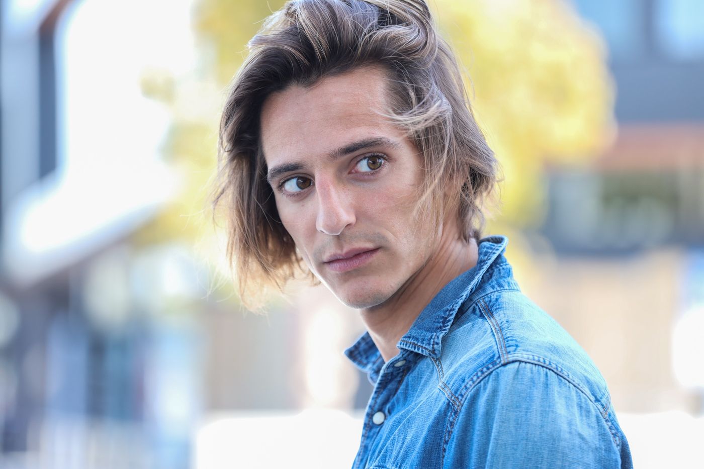 Matteo Leoni con camisa vaquera y el pelo sobre la cara mira hacia atrás por encima del hombro, con una calle desenfocada al fondo.