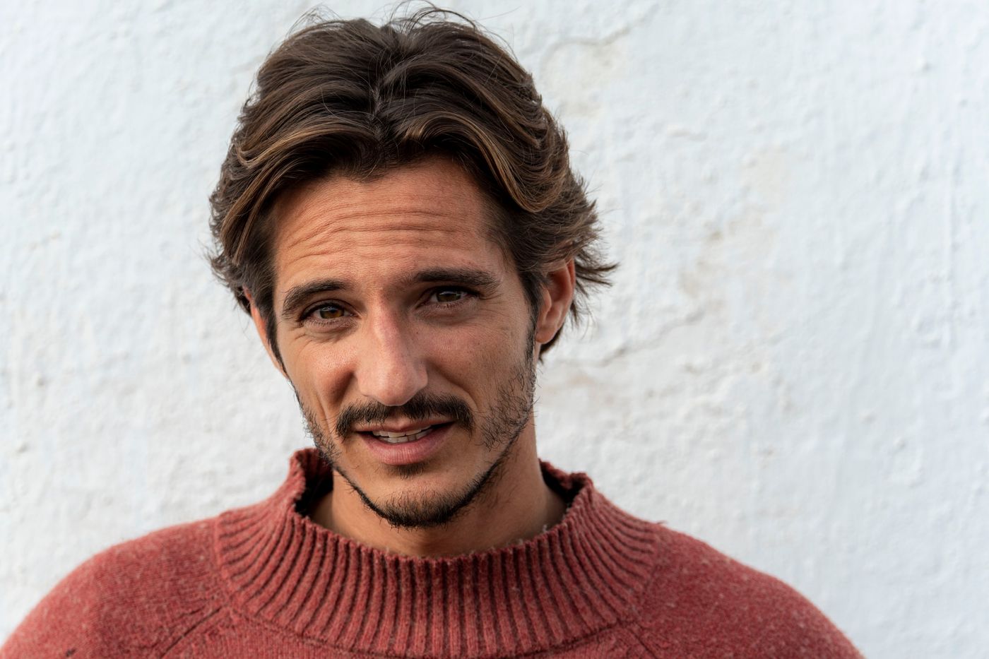 Matteo Leoni sonríe con la boca entreabierta, el pelo ondulado suelto, con un jersey rojo teja delante de una pared blanca.