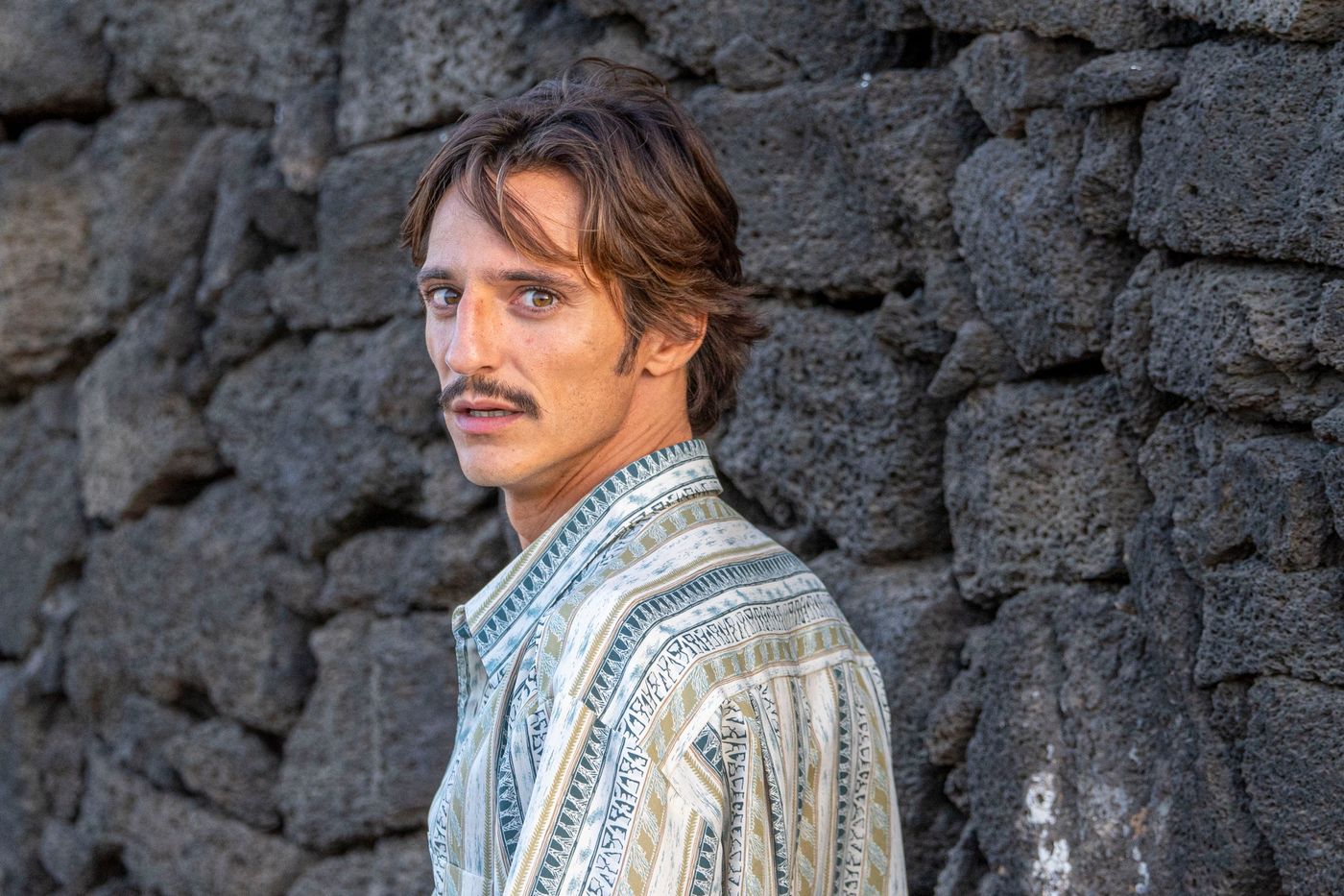 Matteo Leoni con una camisa estampada, girado hacia un lado y mirando más allá de la cámara, delante de un muro de piedra volcánica oscura.