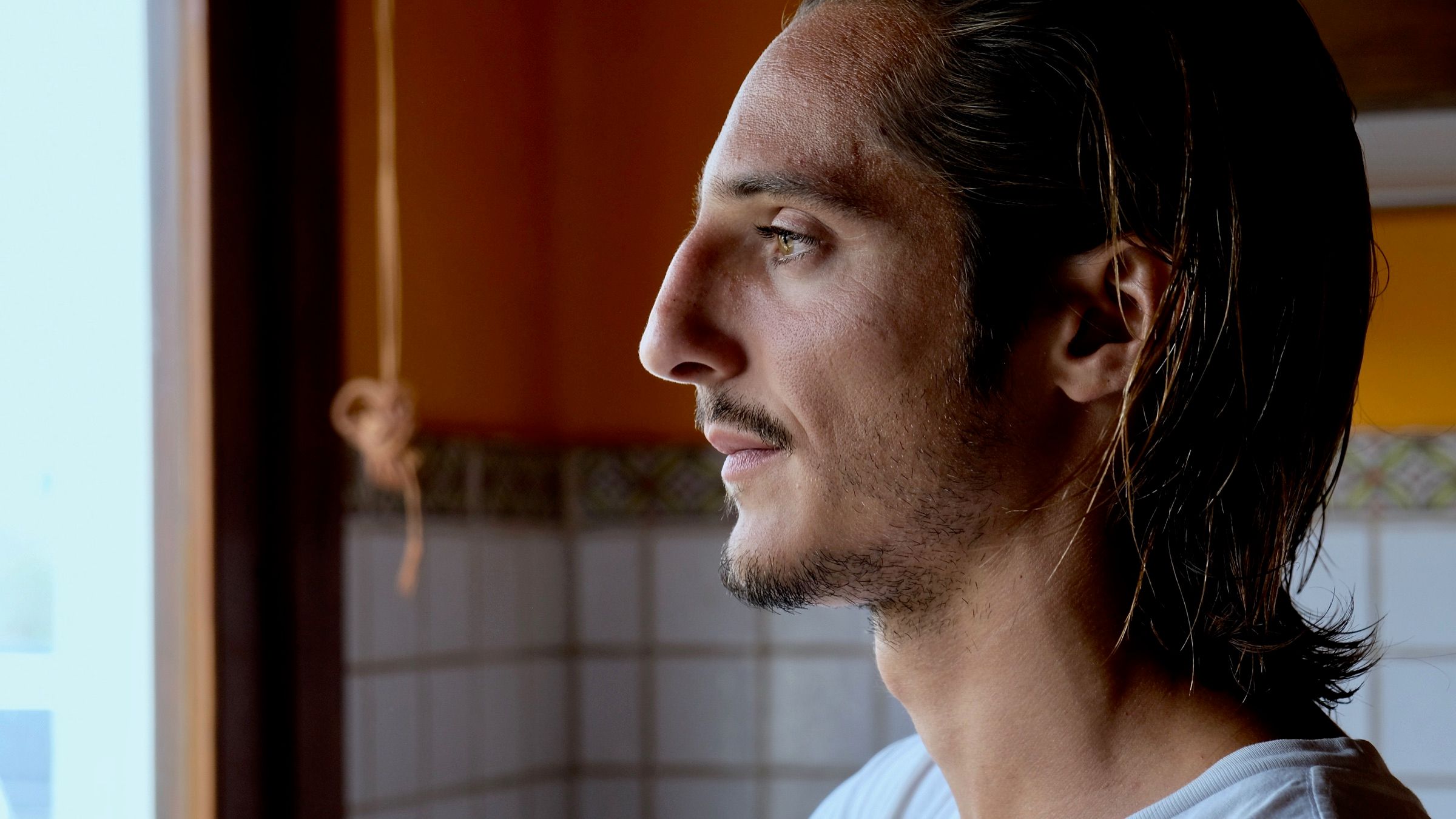 Matteo Leoni de perfil, con el pelo recogido y una camiseta blanca, mira por una ventana en una habitación de paredes con azulejos y luz cálida.