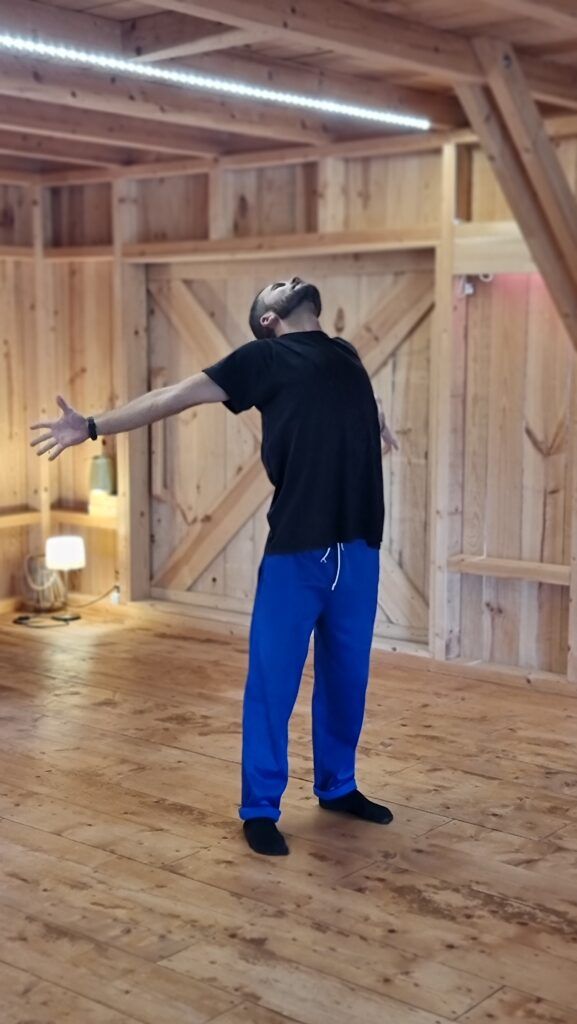 Un participante del curso, en calcetines, de pie en el centro de una sala de madera, con los brazos muy abiertos y la cabeza hacia atrás.