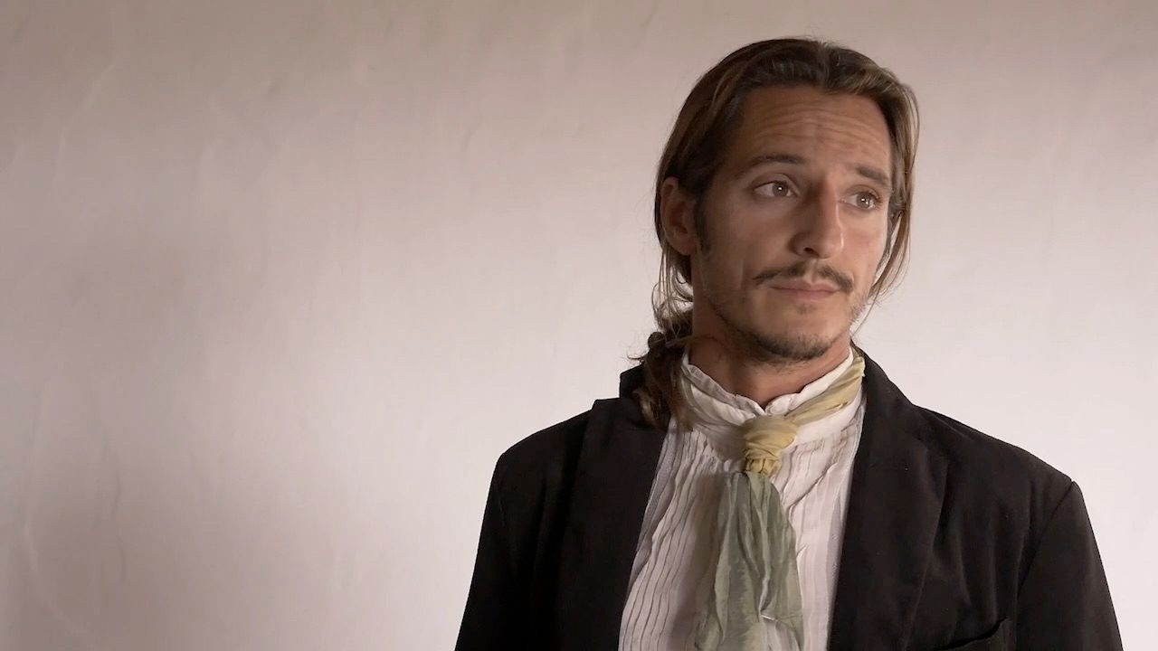 Fotograma de rodaje: un hombre con chaqueta oscura, camisa blanca y un pañuelo verde claro al cuello, el pelo recogido, mirando hacia un lado contra una pared desnuda.